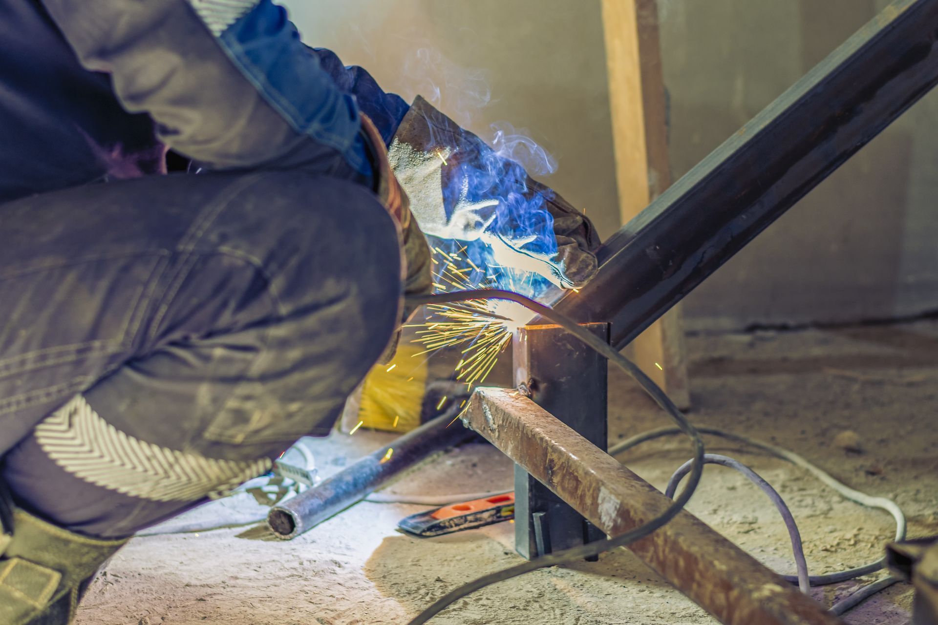 Worker welding a black metal frame on the floor, with blue sparks and tools nearby.