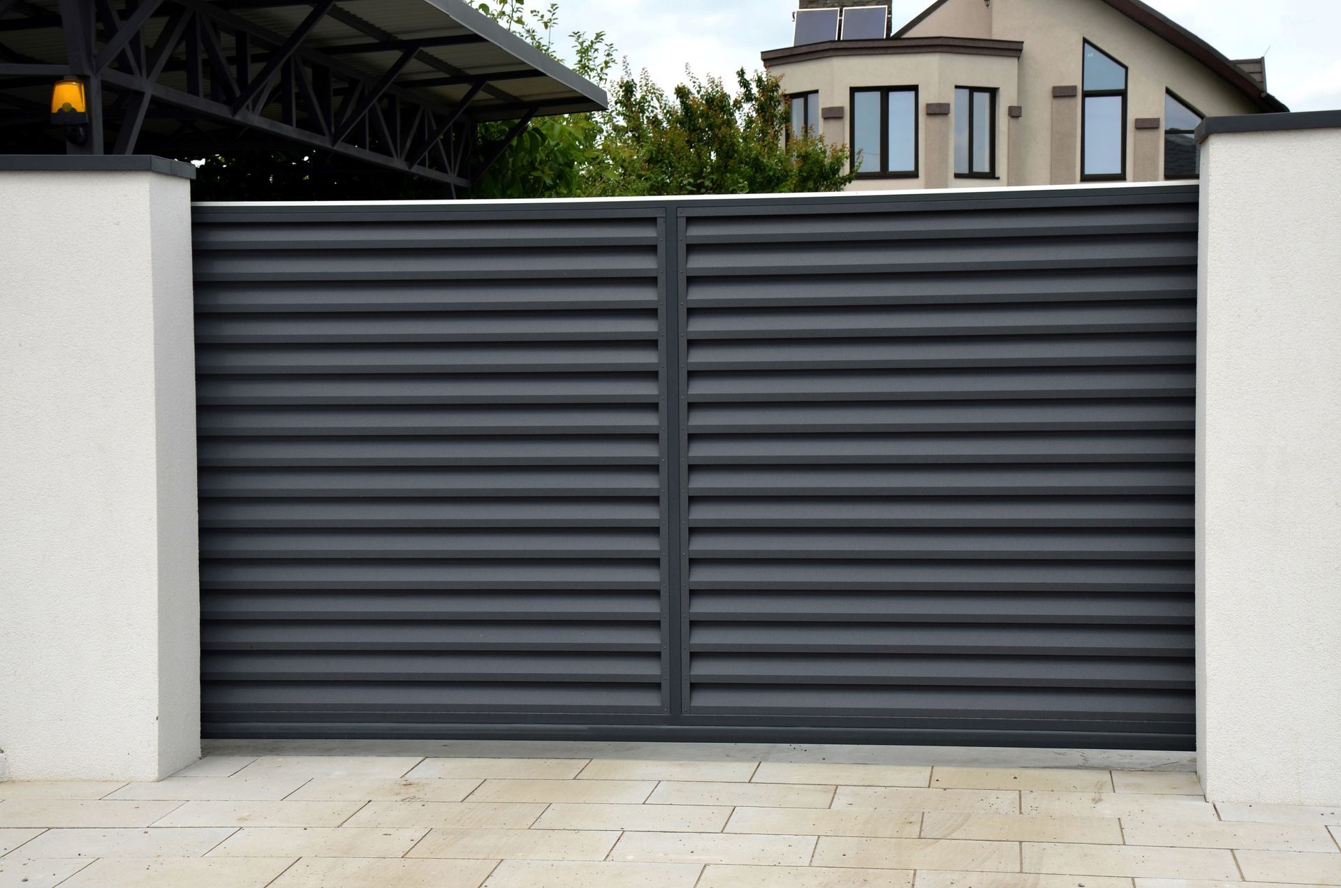 Dark gray metal driveway gate between white pillars in front of a modern house