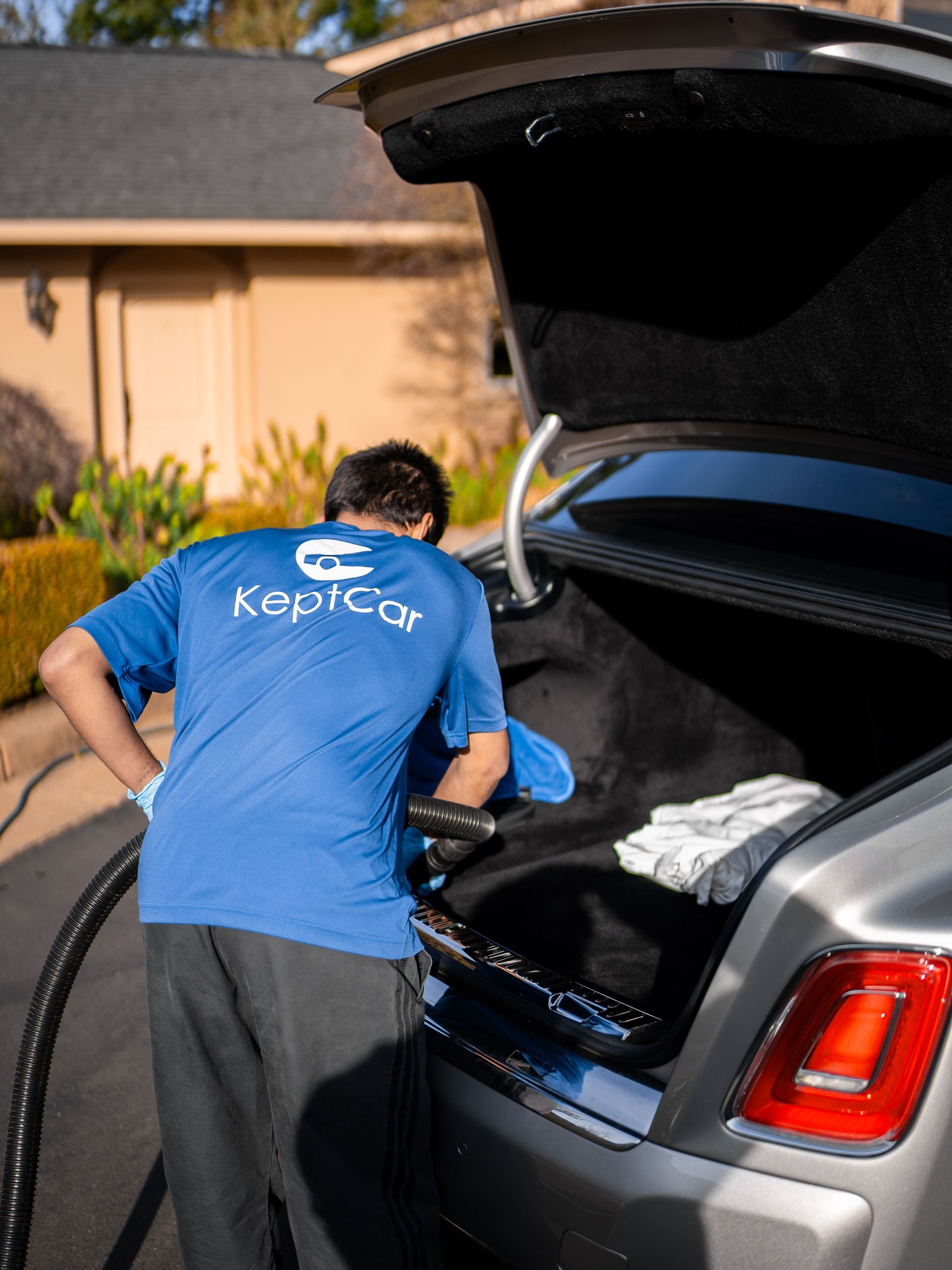 Person in a blue Kept Clean shirt cleaning the open trunk of a silver car in a driveway.