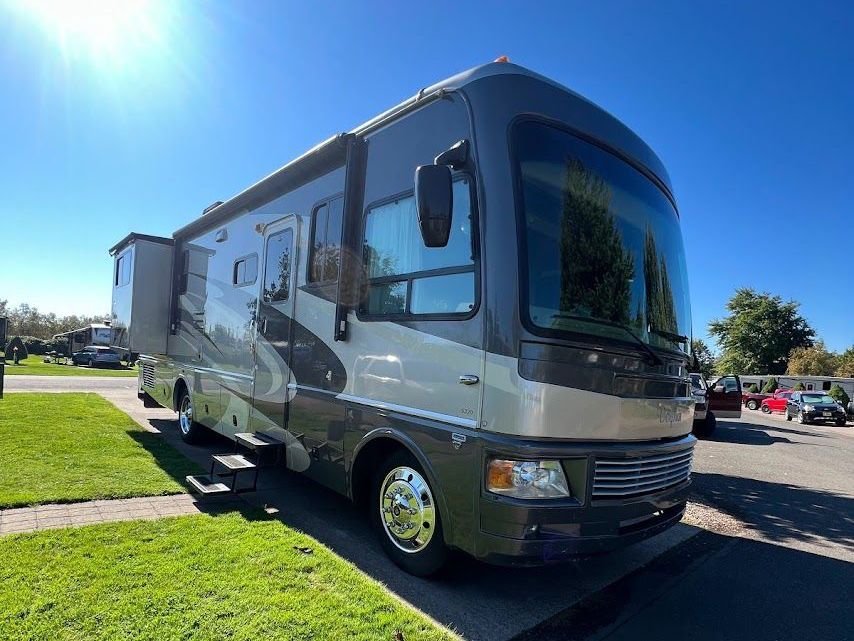 Large gray motorhome parked on grass in a sunny lot, with a clear blue sky overhead.