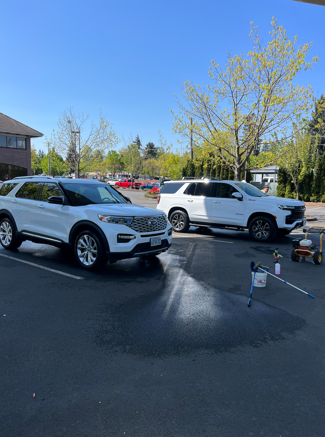 Two white SUVs parked in a wet lot with cones and a spray bottle under a bright blue sky
