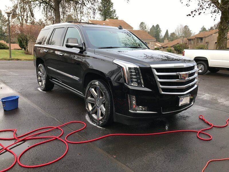 A black cadillac escalade is parked in a driveway next to a red hose getting detailed by keptcar