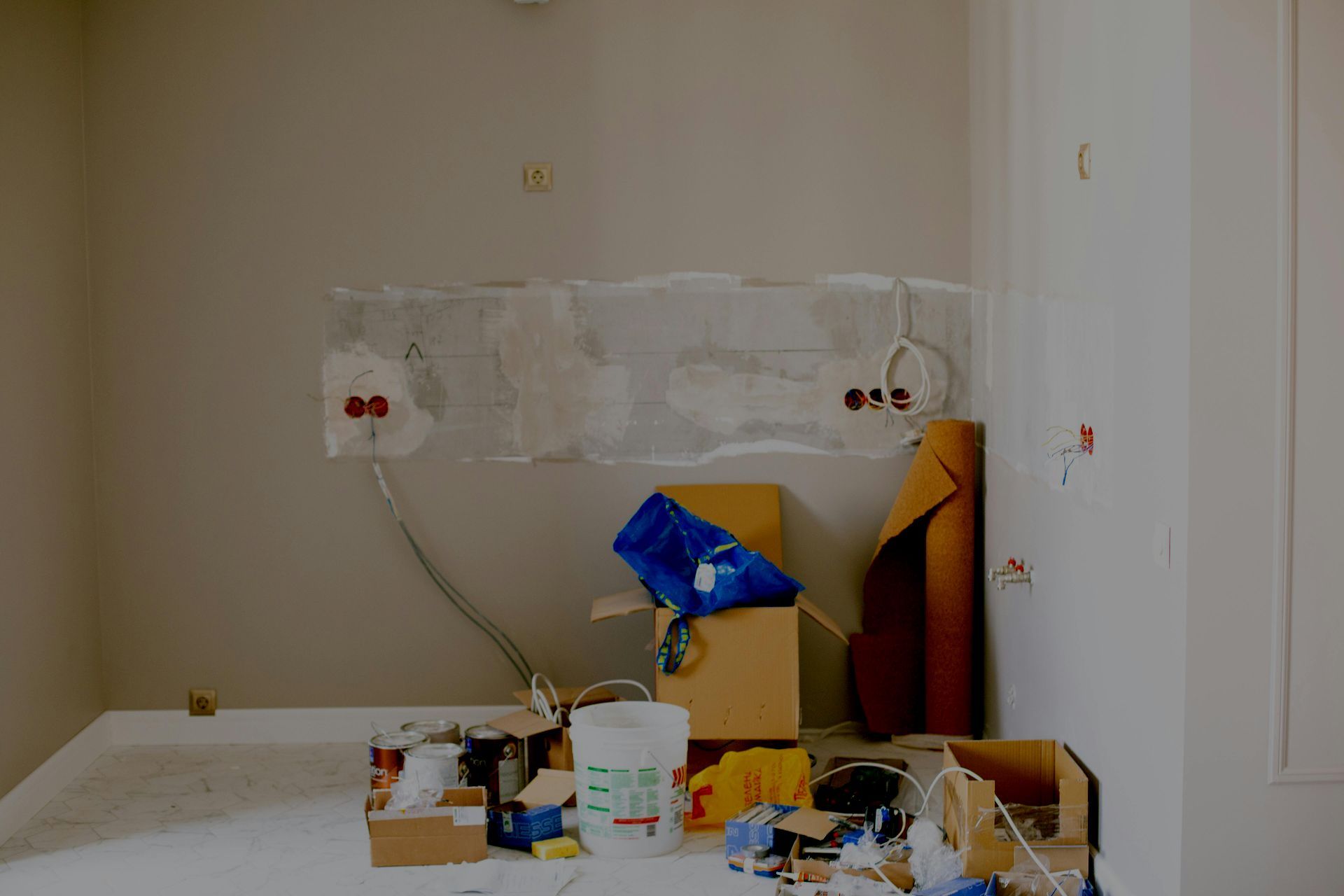 Cluttered corner with a table, boxes, and tools against a wall with outlets and taped plastic sheets