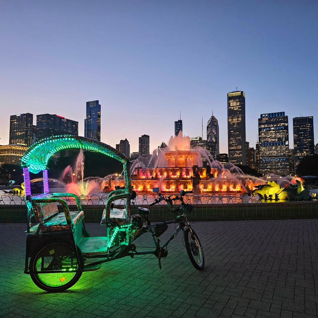 An evening pedicab city tour featuring an illuminated chicago rikshaw ride parked before the glowing Buckingham Fountain and skyline.