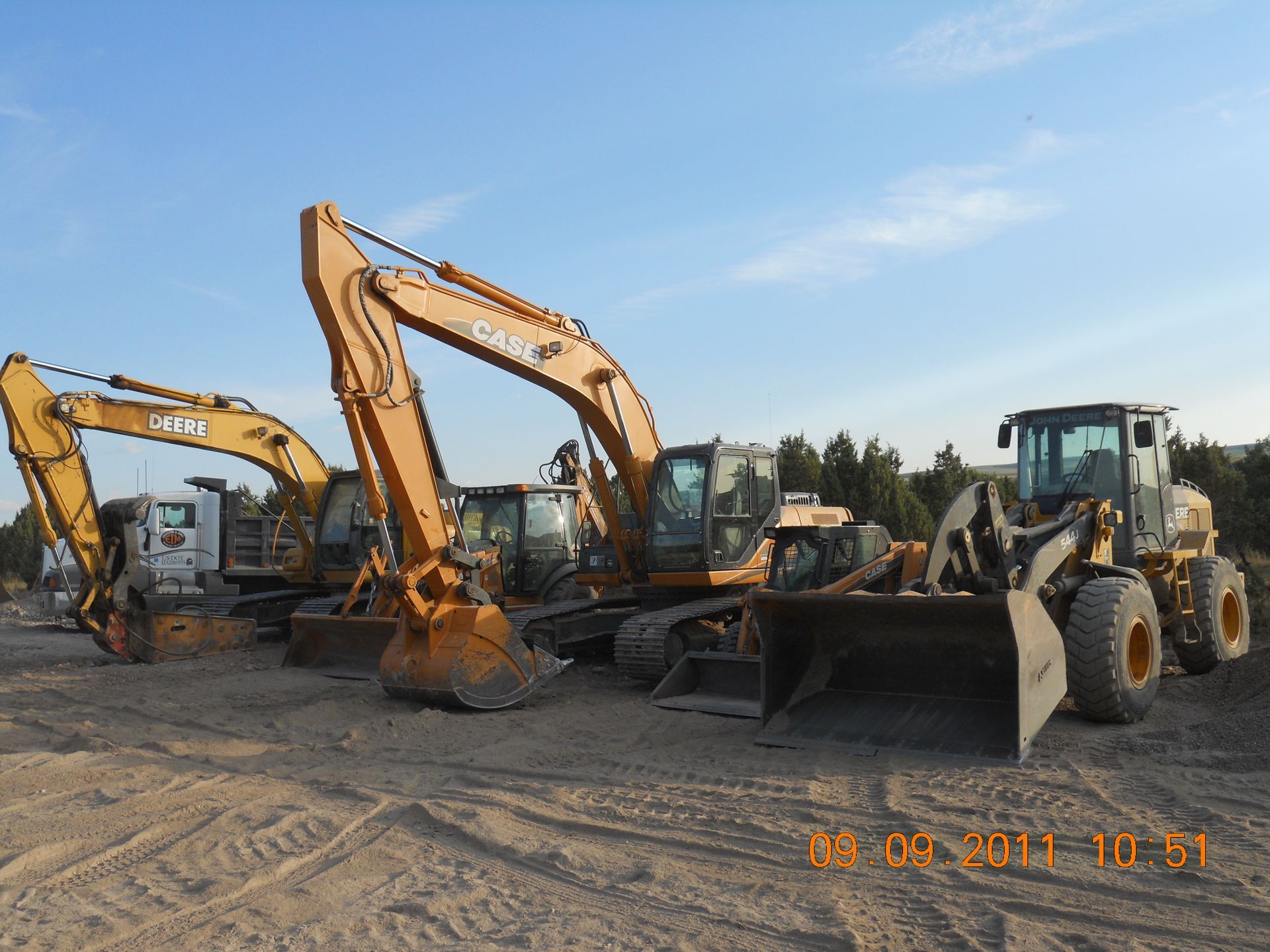 Construction equipment: excavators and a loader parked on a dirt lot under a blue sky.
