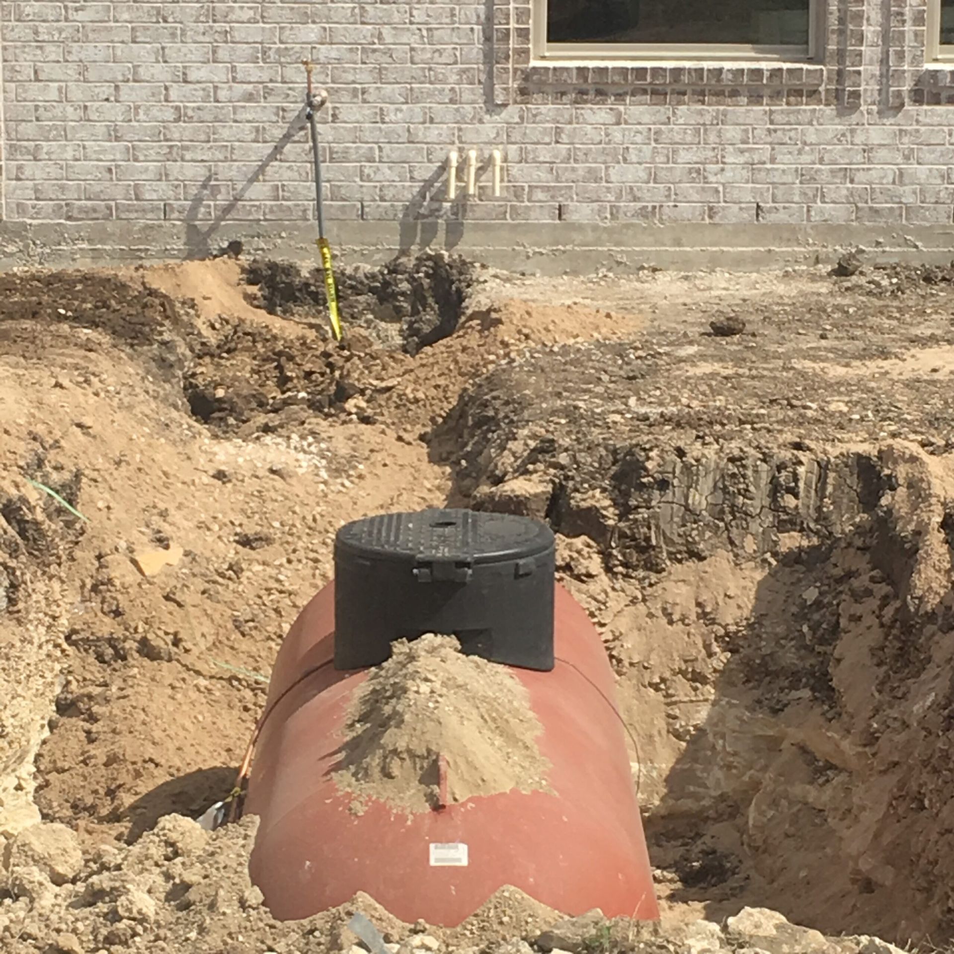 A brown septic tank in a dug trench, with a black lid. Brick wall in the background.