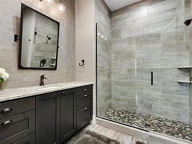 Bathroom with gray tile, dark cabinets, granite countertop, and glass shower with pebble floor.