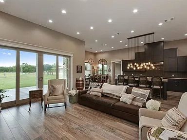 Spacious living room with brown leather sofa, beige chairs, sliding glass doors, and open kitchen with dark cabinets.