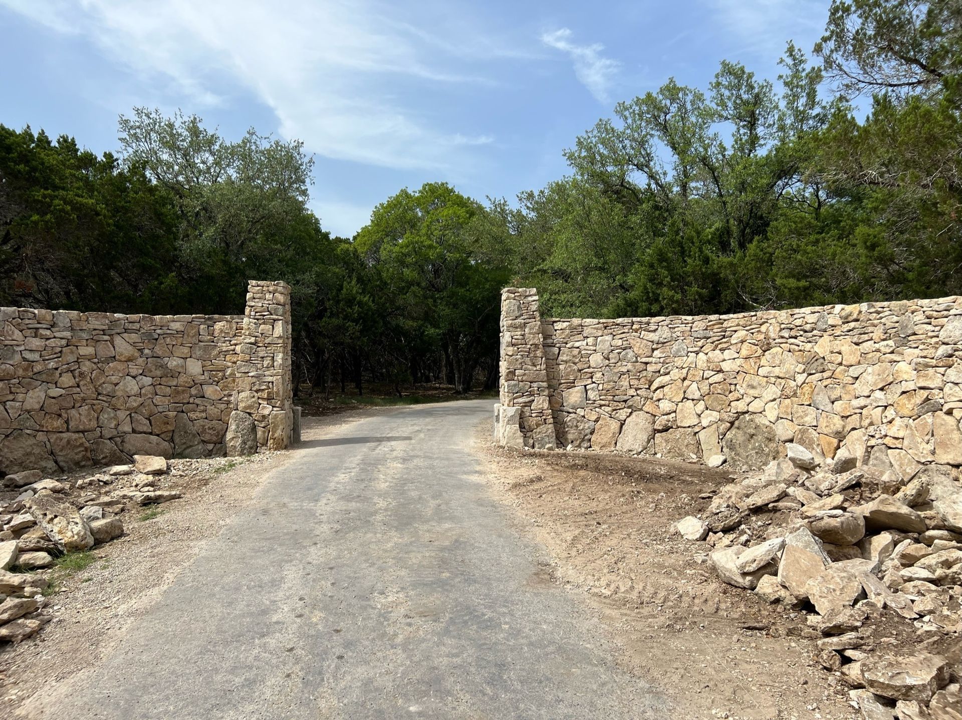Stone walls flank a narrow road leading into a wooded area under a blue sky.