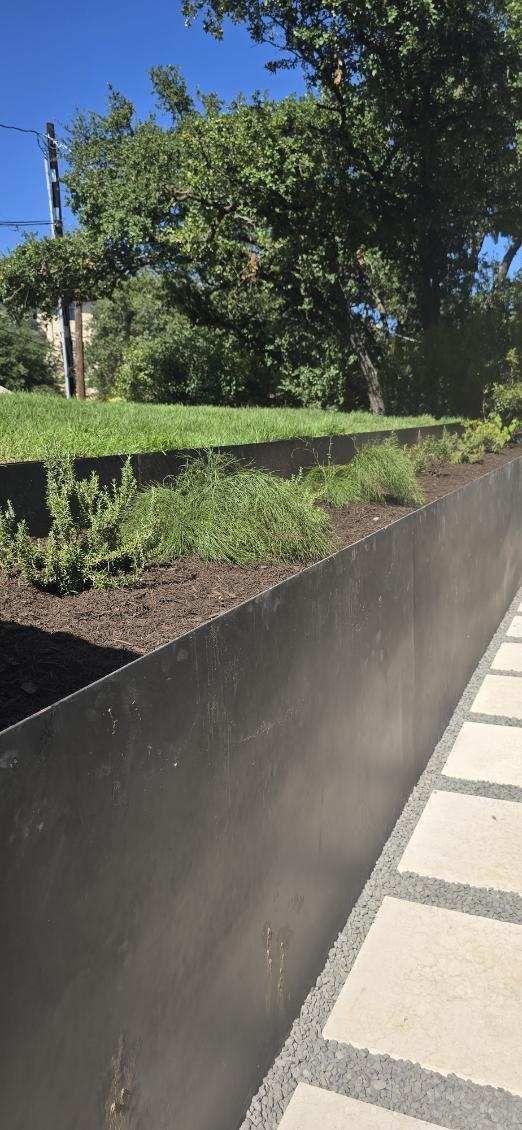 A long, dark metal planter filled with soil and plants, next to a stone walkway and greenery under a blue sky.