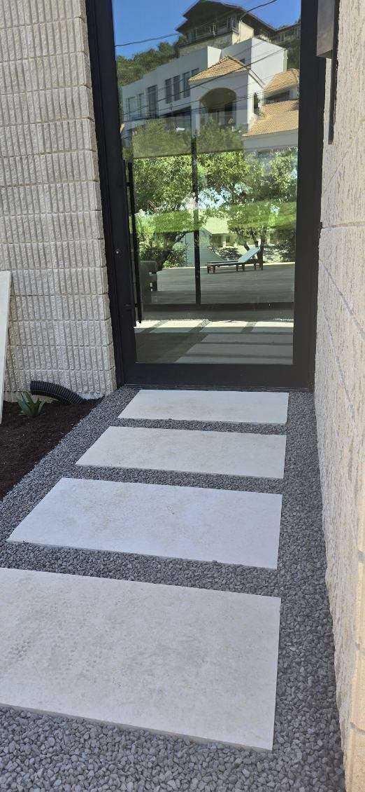 Stone path leading to a doorway, flanked by a brick wall and gravel, with a view of buildings and trees beyond.