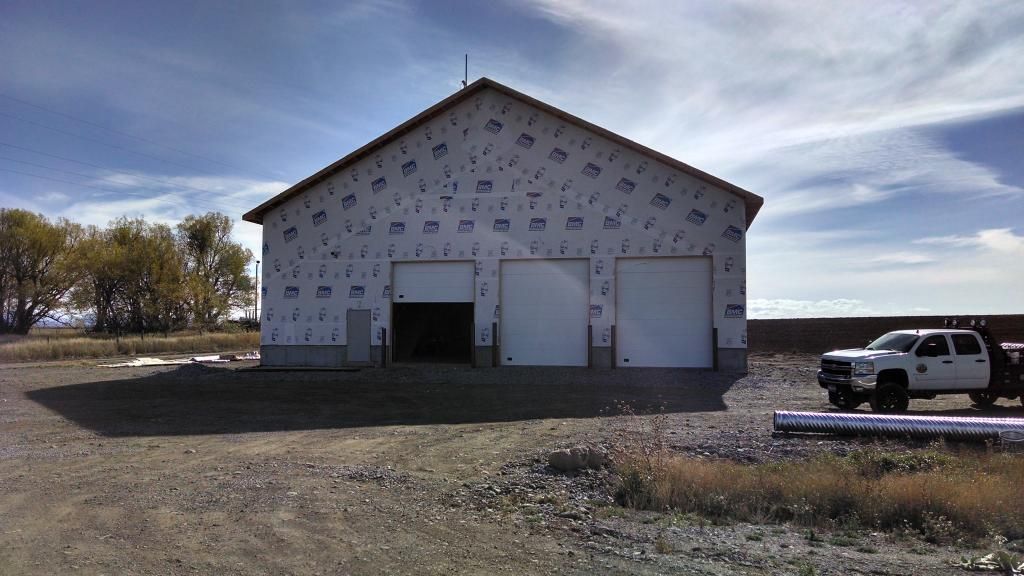 Building with two garage doors, unfinished siding, and a truck parked outside. Sunny day.