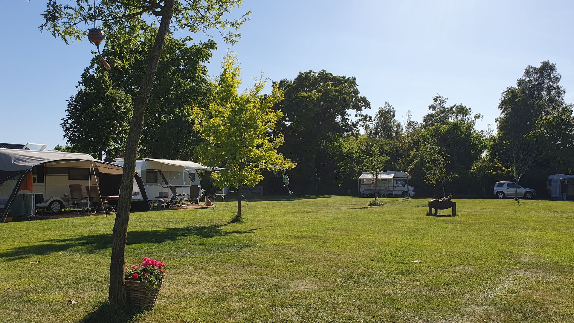 Een campingtafereel met caravans, een grasveld, bomen en een blauwe lucht.