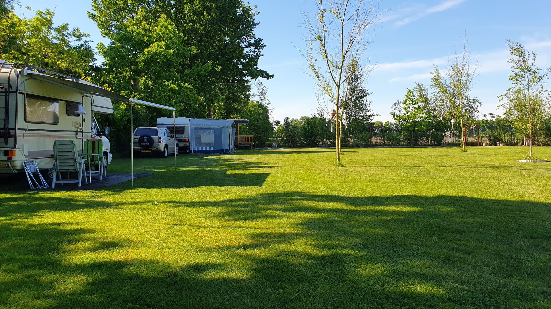 Een camperplaats met een groen gazon, voertuigen, bomen en een blauwe lucht.