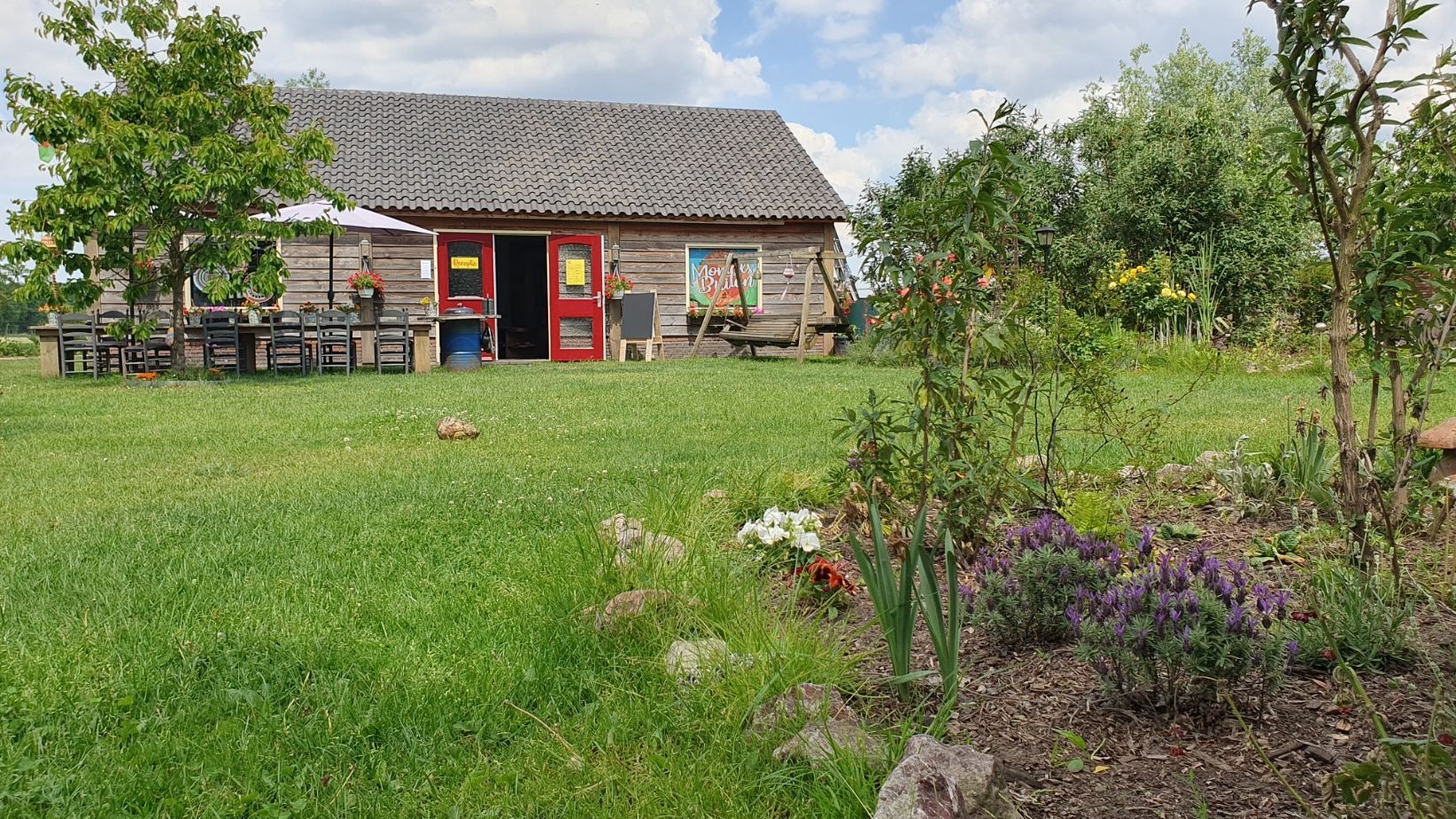 De kantine en receptie met rode deuren op een grasveld. Een bloembed op de voorgrond, bomen en tafels op de achtergrond.