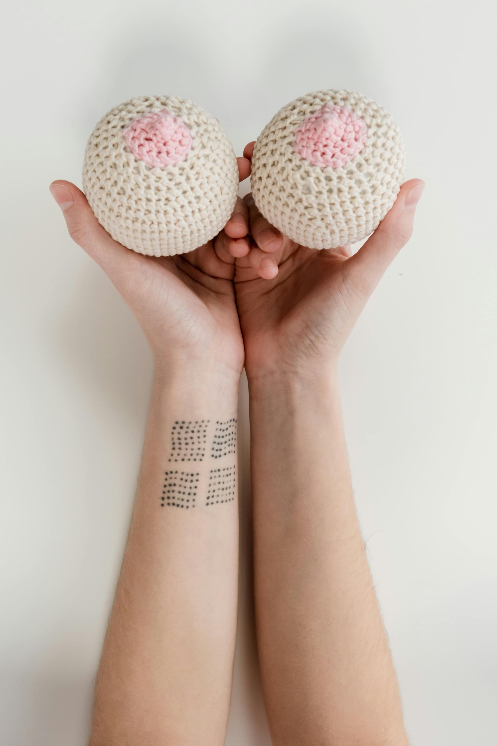 Handen die twee gehaakte borsten met roze tepels vasthouden; tatoeage op de arm.