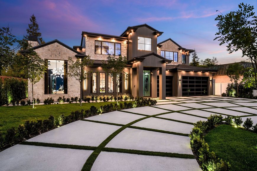 Two-story luxury home with stone and board-and-batten exterior geometric paver driveway and exterior lighting at dusk.