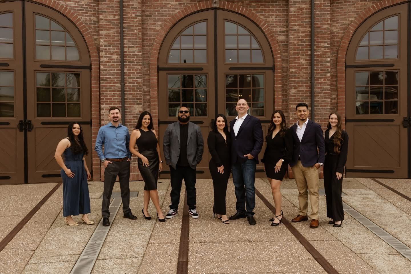 Group of nine real estate professionals posing in business attire in front of a brick building with arched windows.