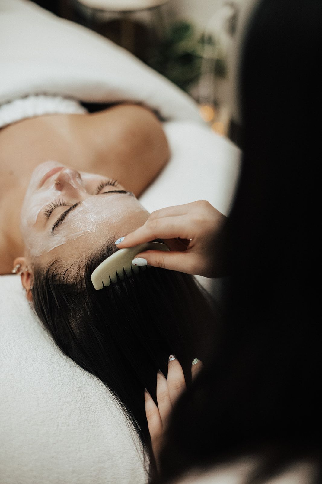 Woman receiving a scalp massage with a comb while lying on a spa bed, attended by someone in a black shirt.