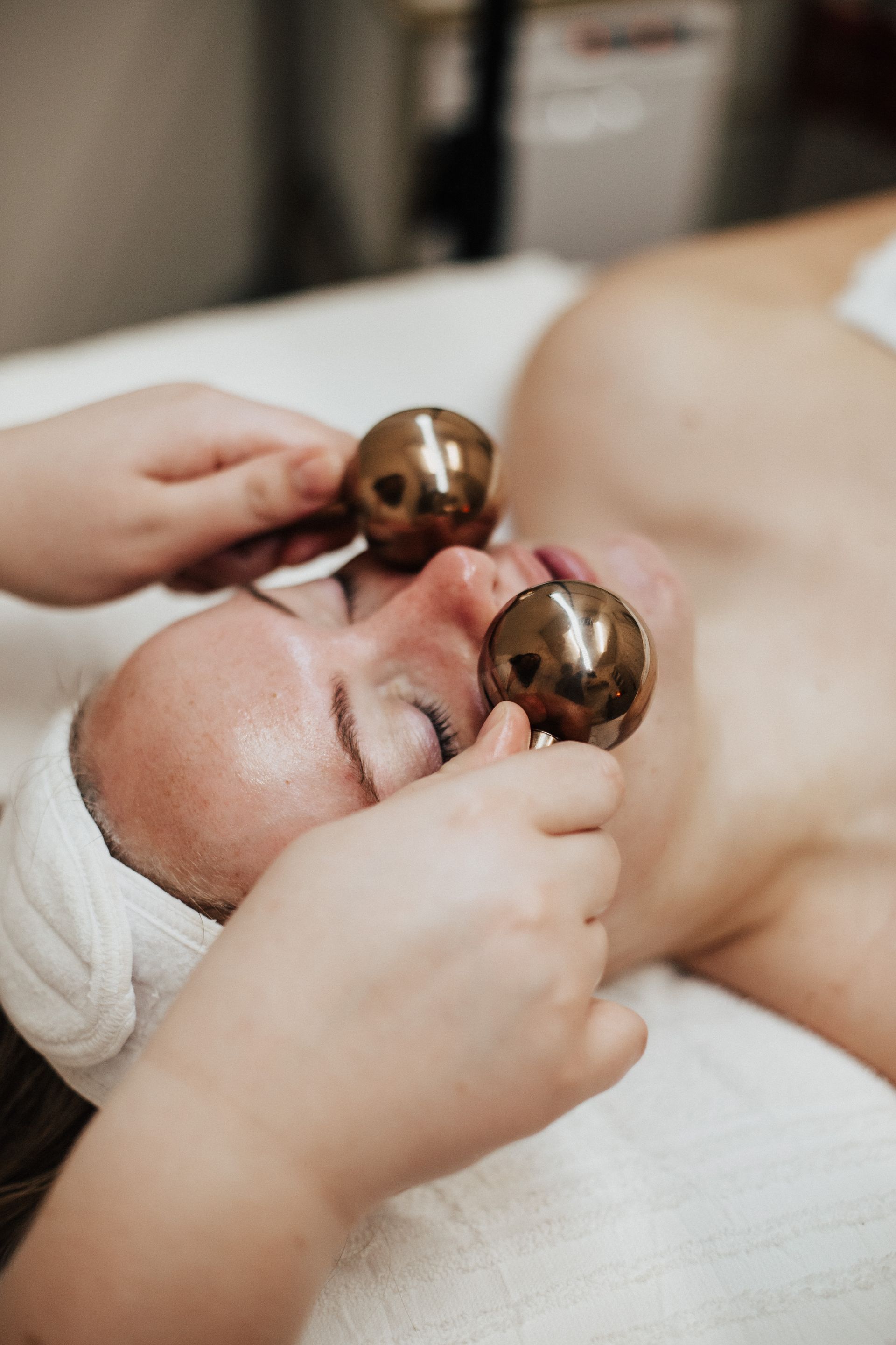 Person receiving facial treatment with metal globes on eyes, lying on a spa table.