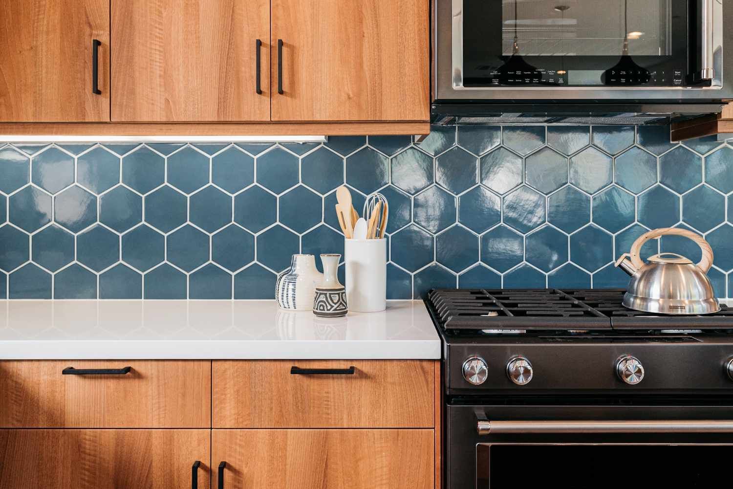 Blue hex-tile kitchen backsplash with wooden cabinets, white counter, and black stove with kettle