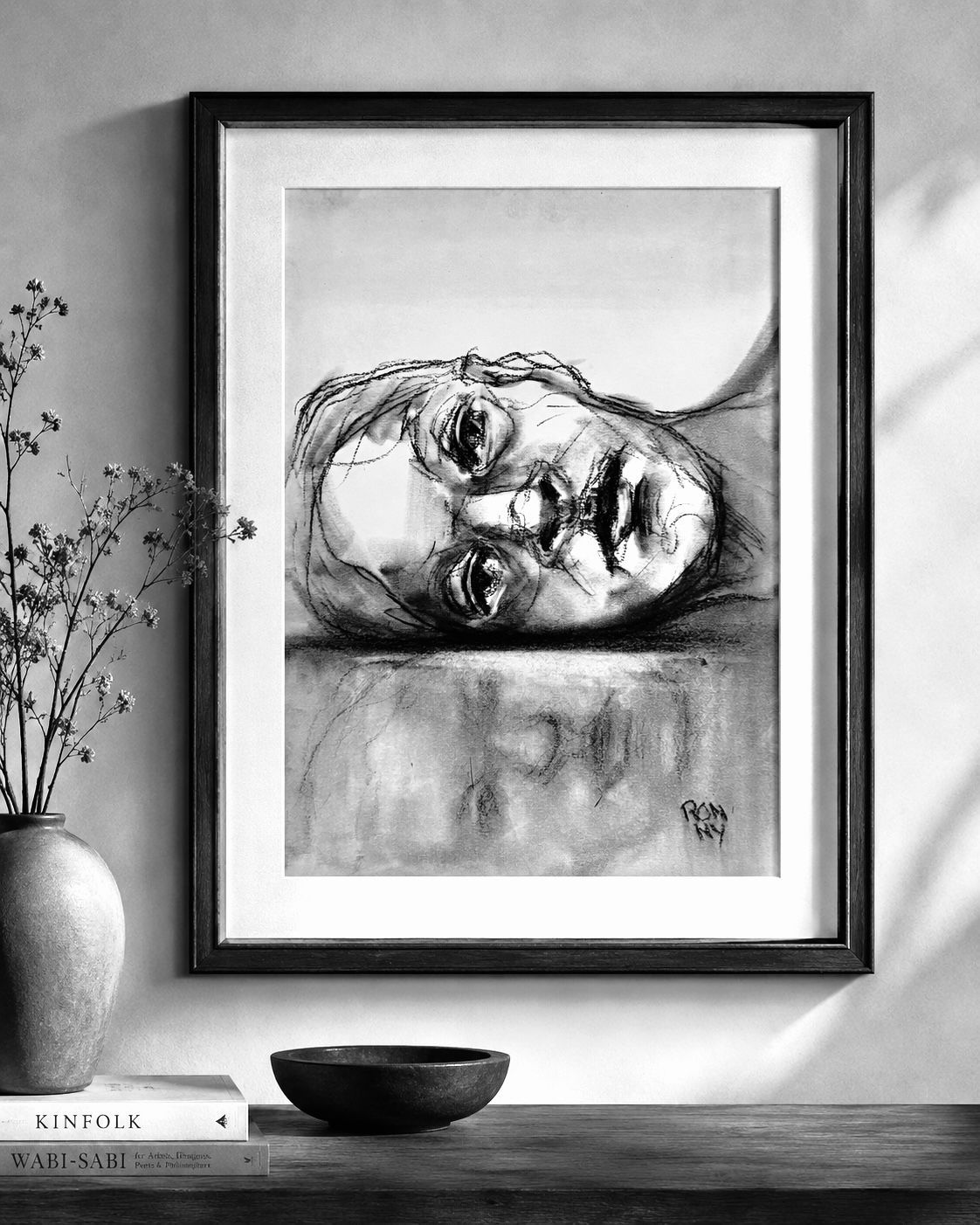 Framed charcoal portrait of a reclining face on a wall above a shelf with vase and bowl