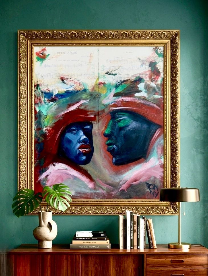 Kleurrijk abstract portret boven een houten dressoir, met lamp, boeken en een kleine vaas in een turquoise kamer.