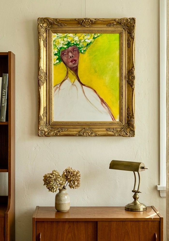 Ingelijst abstract portret boven een houten dressoir met bloemen en een messing lamp in een lichte kamer.