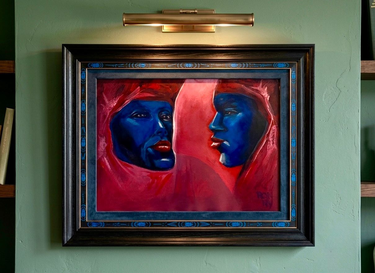Ingelijst schilderij van twee blauwe gezichten tegen een rode achtergrond op een groene muur.