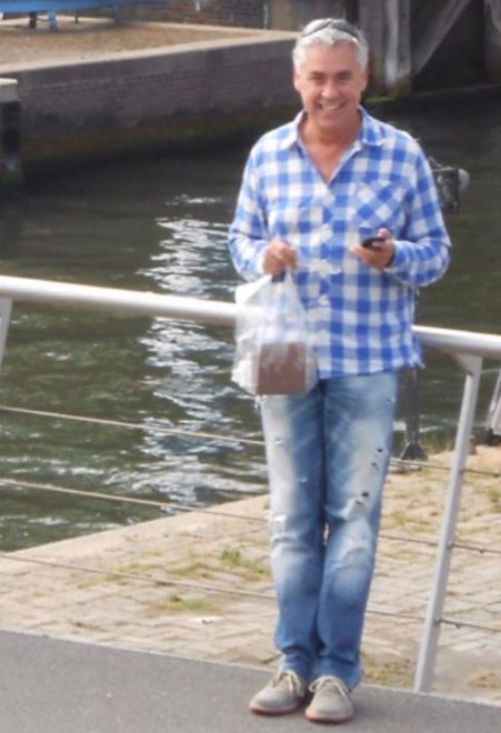 Een persoon in een blauw geruit overhemd staat bij een kanaal en houdt een telefoon en een plastic tas vast.