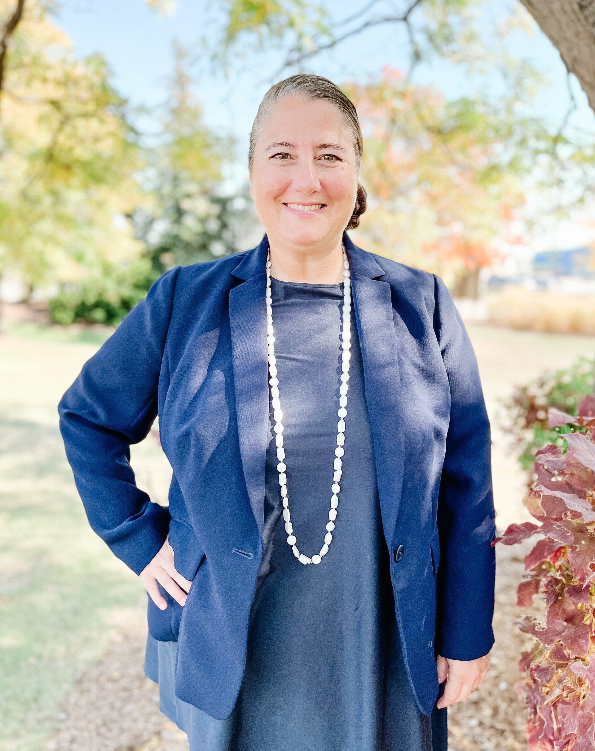 A woman wearing a blue jacket and a long necklace is standing in front of a tree.