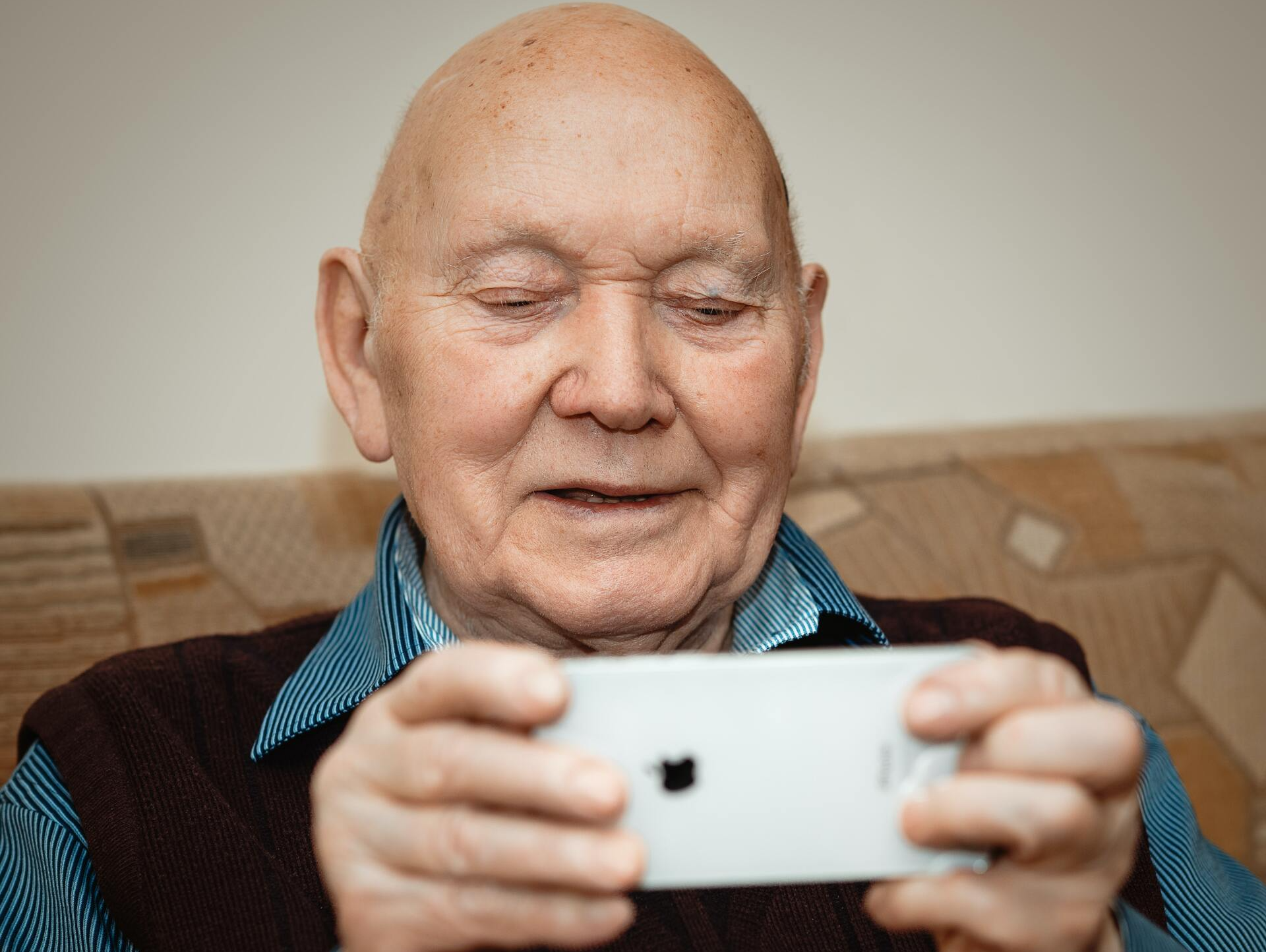 An elderly man is sitting on a couch looking at a cell phone.