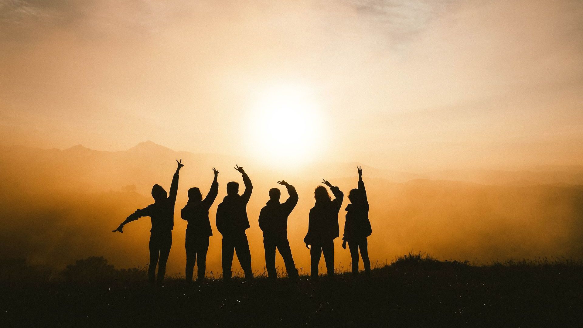 A group of people are standing in front of a sunset with their arms in the air.