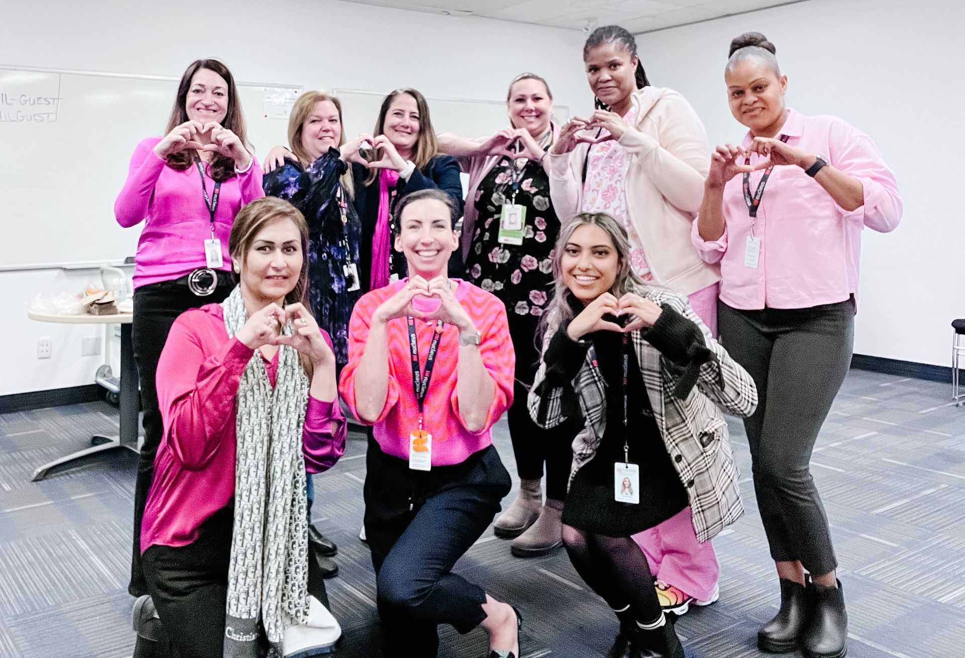 A group of women in pink shirts are making a heart shape with their hands.