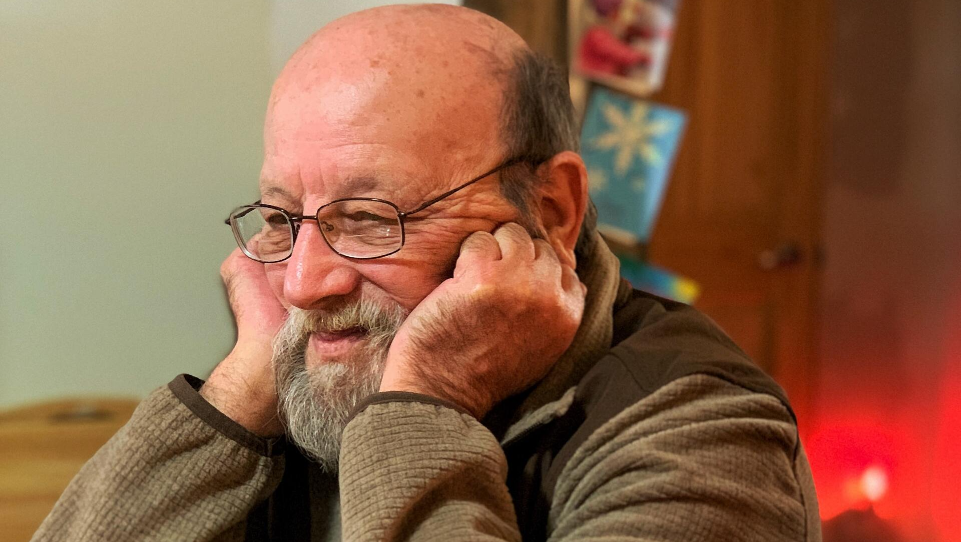 A bald man with a beard and glasses is sitting at a table with his hands on his face.