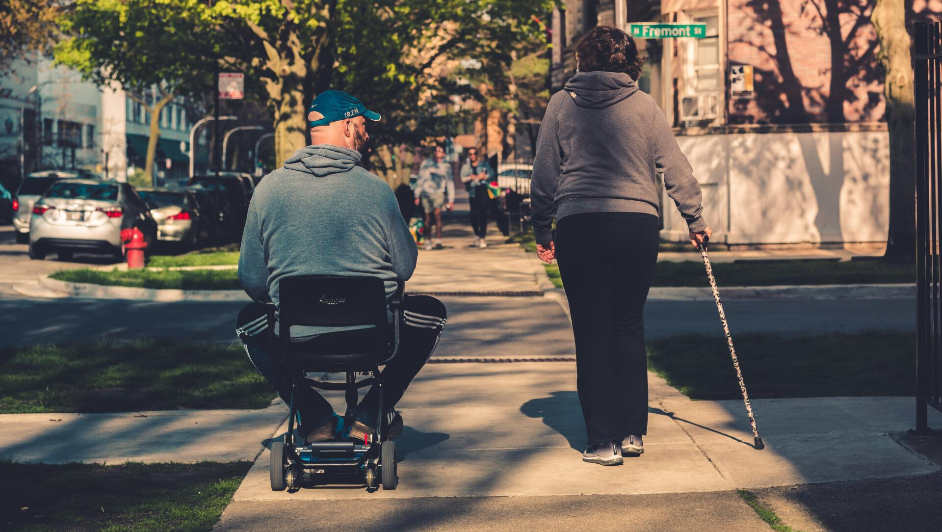 A man in a wheelchair and a woman with canes are walking down a sidewalk.