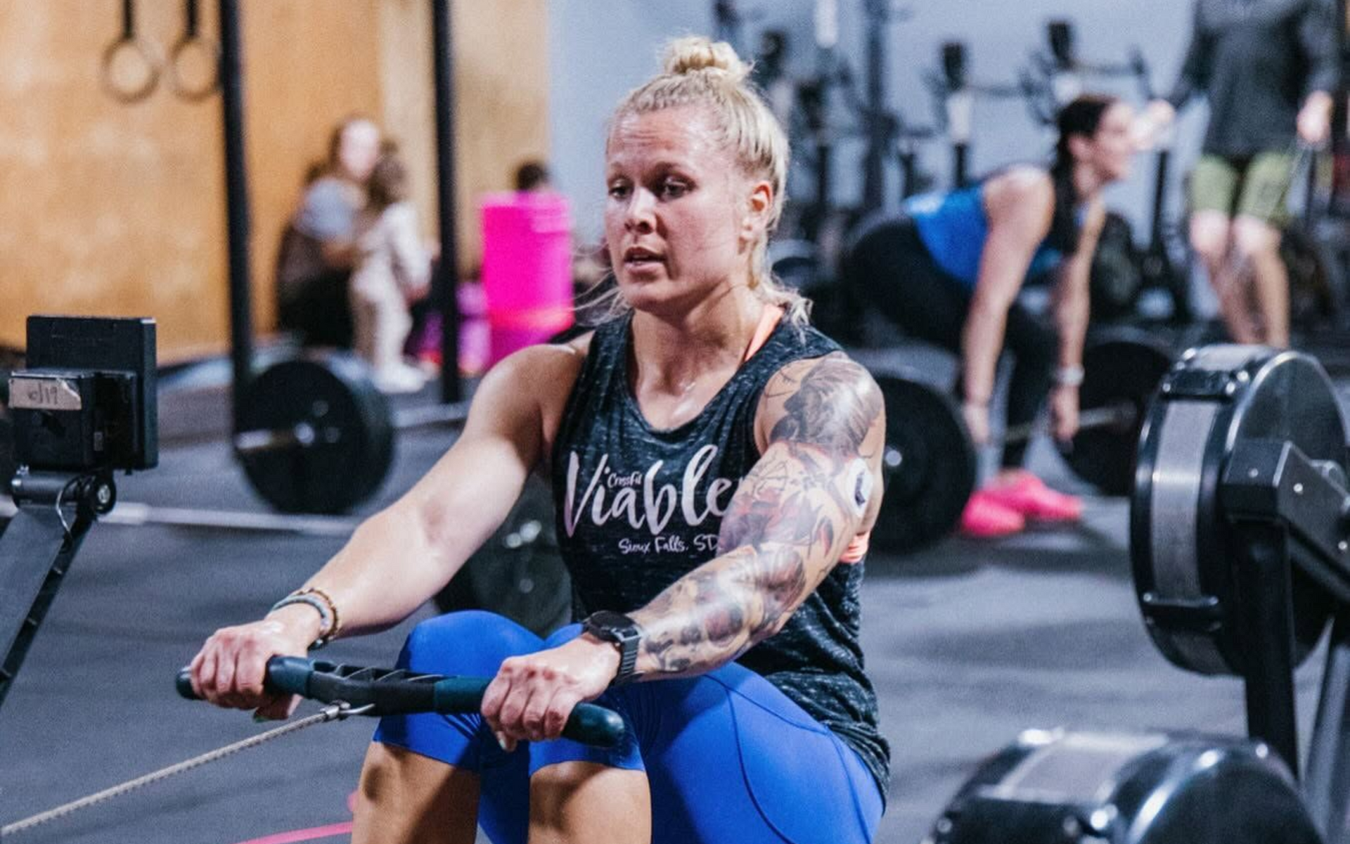 Woman kneeling and writing on a gym floor during a workout, with another woman stretching beside her.