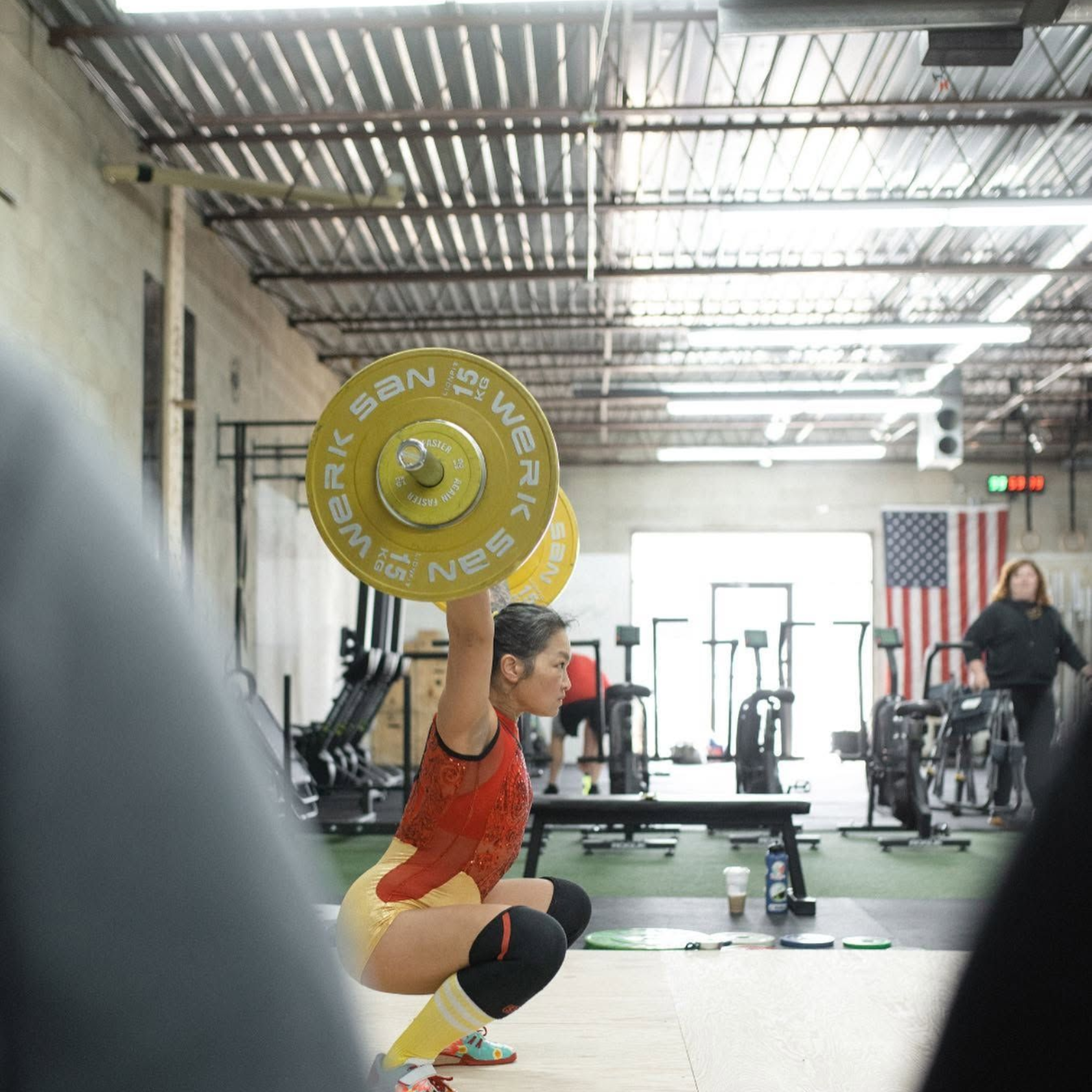 Athlete squatting with a yellow barbell in a gym, wearing an orange top and black knee sleeves at the best CrossFit gym in Sioux Falls, SD.
