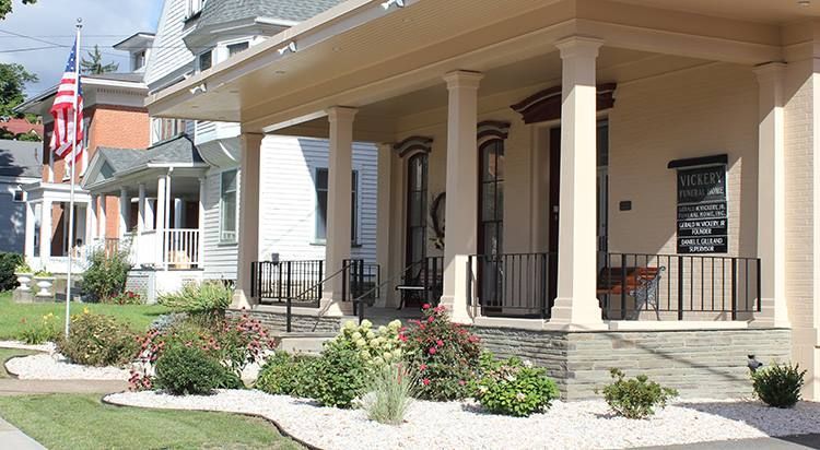 Row of historic houses, one with a large porch supported by white columns. Flowers and shrubs in the foreground.