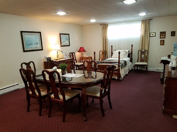 A bedroom with a dining table, bed, and dressers. Red carpet and white walls. Contains artwork and a window with cream-colored curtains.
