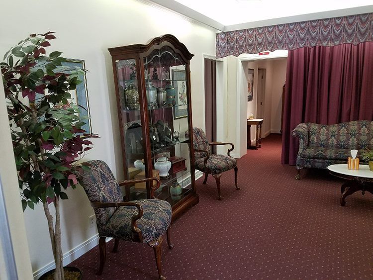 A waiting area with burgundy carpet, floral patterned chairs, and a display cabinet. A sofa and doorway with red curtains are visible.