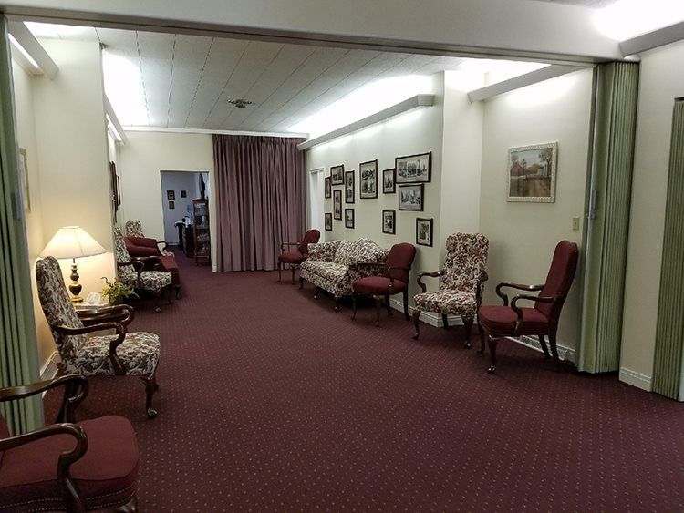 Awaiting room with maroon carpet, floral chairs, and a maroon couch. The walls are cream-colored with framed photos and a doorway.