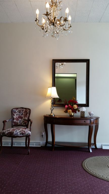 An elegant room with a chandelier, mirror, table with a lamp, floral arrangement, and upholstered chair.