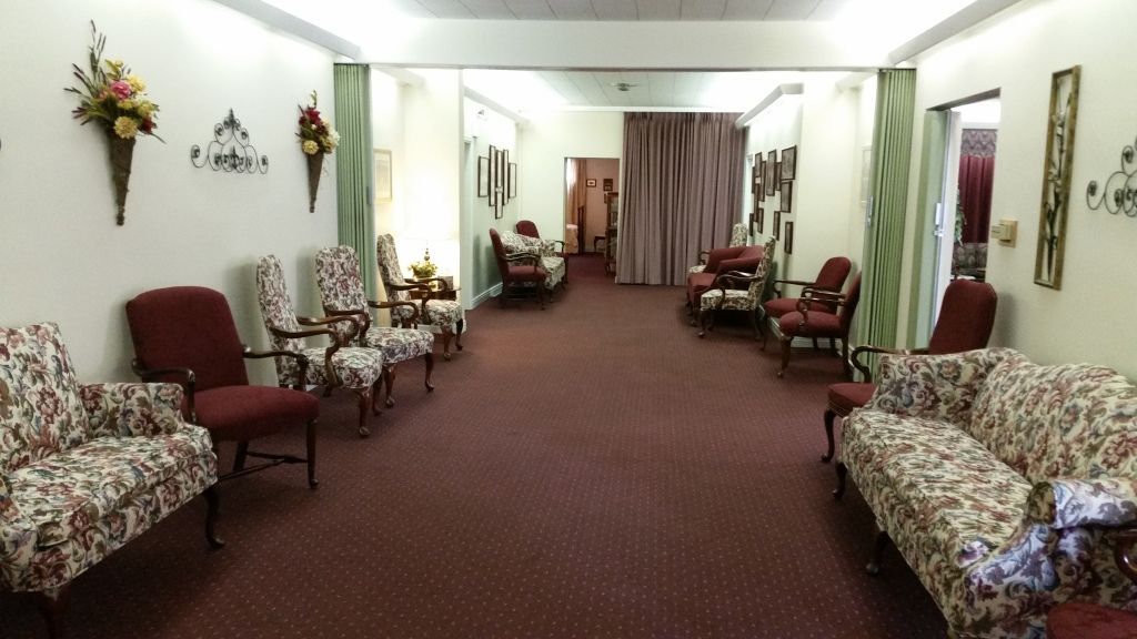 Long, formal living room with red carpet, patterned couches, and several ornate chairs. Walls are white, with floral wall decorations.