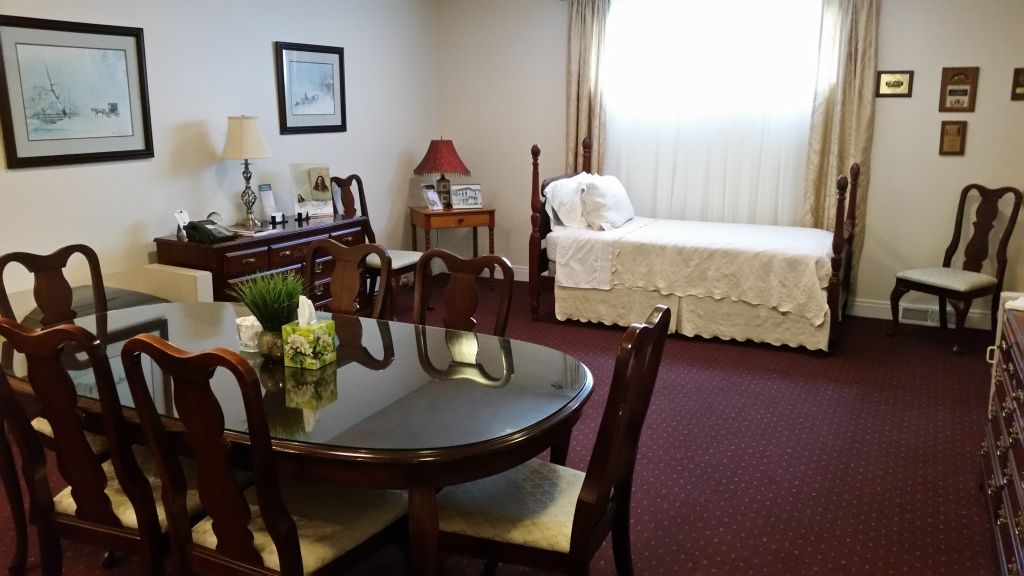 A furnished room with a bed, dining table, and chairs. The room has artwork, lamps, and a window with curtains, all with a red carpet.