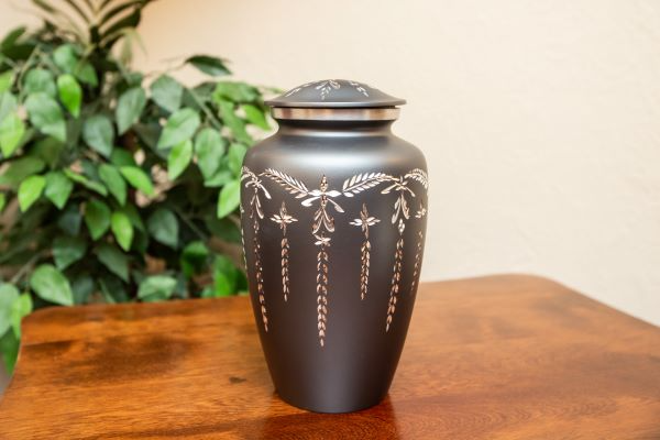 Black urn with etched silver floral design, sitting on a wooden table with a leafy plant background.