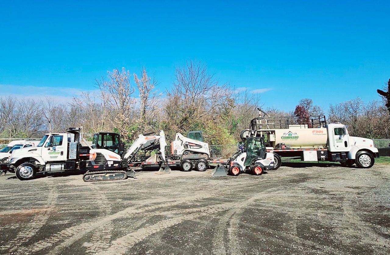 A row of landscaping trucks and equipment parked on a dirt lot beneath a blue sky.