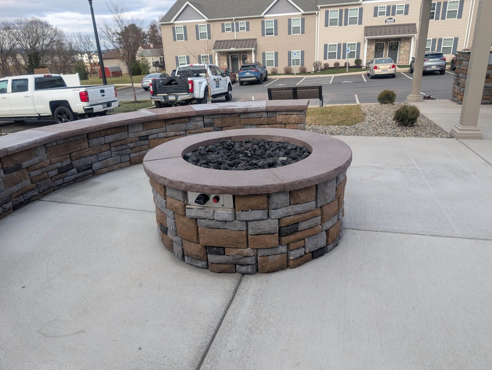 Fire pit on a concrete patio with a surrounding low brick wall in front of an apartment building.