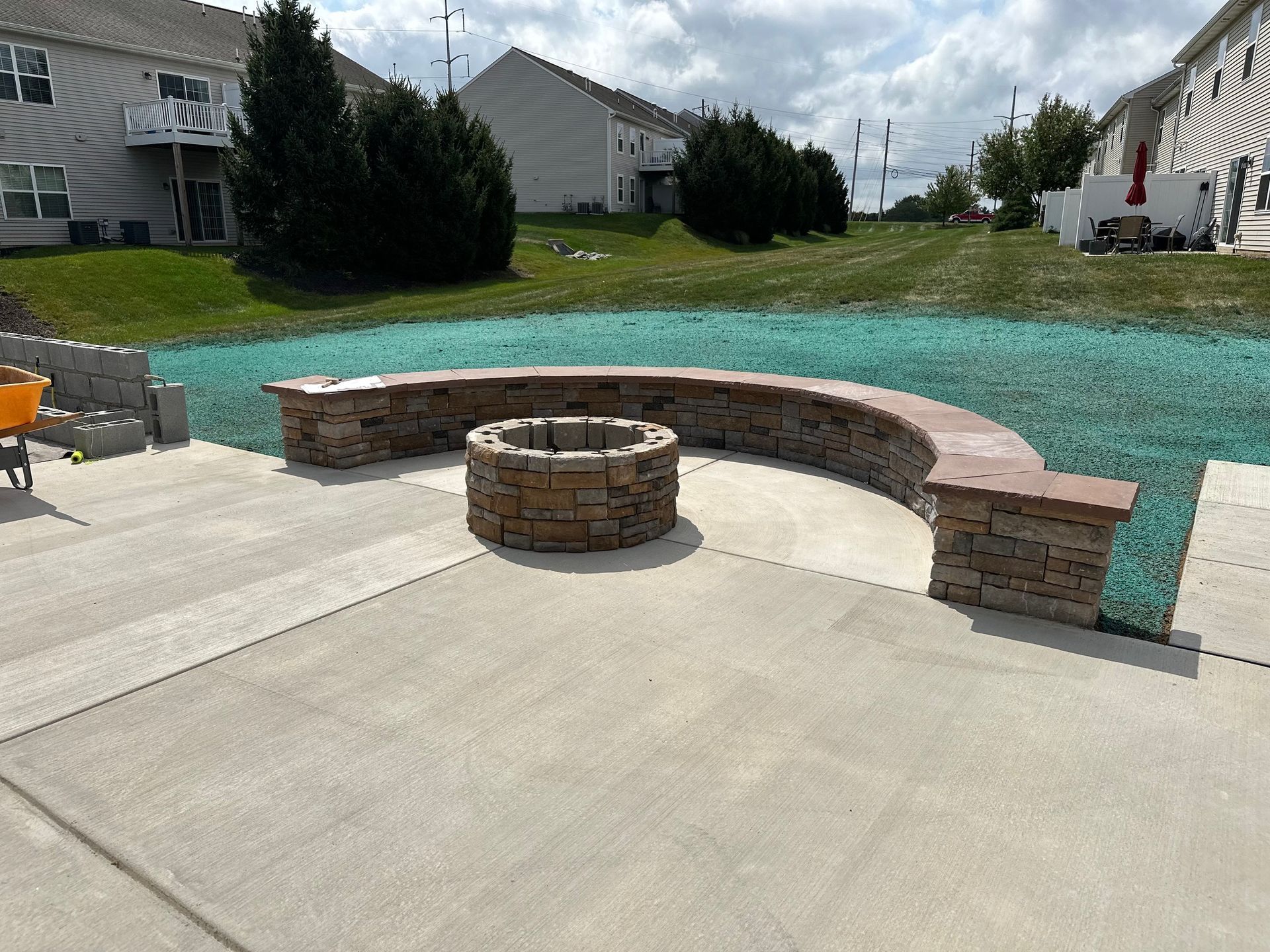 Curved stone fire pit and seating area on a concrete patio, surrounded by grass and houses.