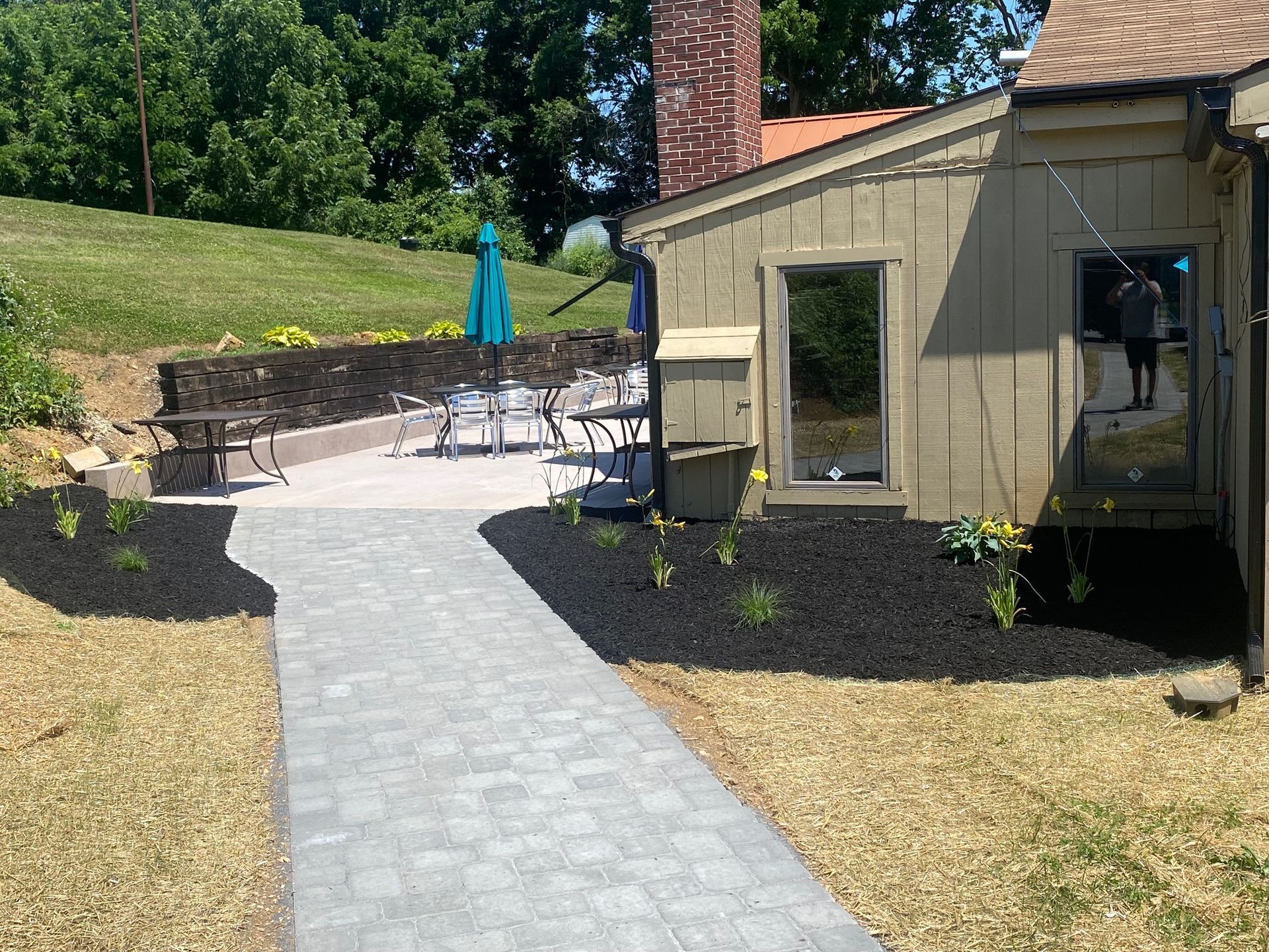 A paved walkway leads to a patio with seating and a building; dark mulch borders the walkway.